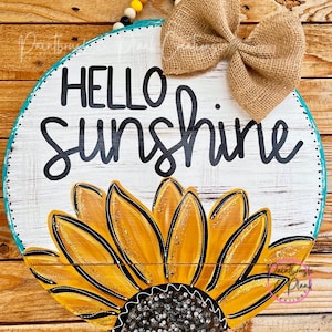 May include: Round wooden sign with a sunflower design and the words "HELLO SUNSHINE". The sign has a burlap bow and a beaded hanger. The sunflower is yellow and black.
