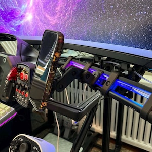 May include: A black and silver gaming setup featuring a phone holder, joystick, and a large monitor displaying a purple and blue nebula. The setup includes a black metal frame and various control panels.