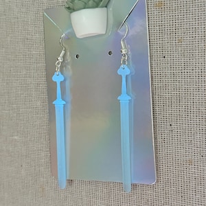 Puede incluir: Pendientes en forma de espada azul claro con ganchos plateados. Los pendientes se exhiben en una tarjeta holográfica con una pequeña decoración de suculenta en la parte superior. El fondo es una tela texturizada de color neutro.