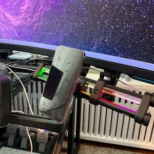 May include: A black phone is mounted on a black and clear device with colorful lights. The device is attached to a black frame, with a large monitor displaying a purple and white galaxy in the background. The setup appears to be for a gaming or simulation setup.