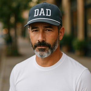Op de afbeelding: Een marineblauwe baseballcap met het woord "DAD" in witte blokletters. De cap heeft een wit touwdetail langs de rand. De persoon draagt een wit t-shirt.