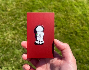 May include: A red rectangular pin featuring a cartoon character with a white body, black hair, and a green gem. The pin is held in a hand against a backdrop of green grass.