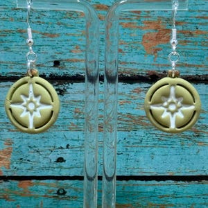 Puede incluir: Un par de pendientes verde oliva con un diseño de rosa de los vientos blanco. Los pendientes son redondos y cuelgan de ganchos plateados. El fondo es una superficie de madera desgastada de color turquesa.