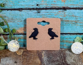 May include: A pair of black raven-shaped stud earrings displayed on a brown earring card. The earrings are set against a backdrop of weathered, turquoise-painted wood. Decorative string lights and greenery are visible.