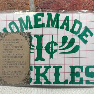 Puede incluir: Una calcomanía verde con las palabras "HOMEMADE PICKLES" en una fuente audaz y estilizada. La calcomanía incluye instrucciones de aplicación y está empaquetada en una funda de plástico transparente. El fondo es una pared de ladrillo rojo.