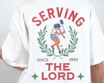 Può includere: T-shirt bianca con grafica a tema tennis. Il design presenta un giocatore di tennis all'interno di una corona d'alloro, le parole "SERVING THE LORD" e l'anno "1995". La maglietta ha una vestibilità ampia e oversize.