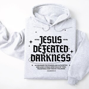 Puede incluir: Una sudadera con capucha gris claro con el texto negro "JESUS DEFEATED DARKNESS", con texto adicional y gráficos de estrellas. La sudadera con capucha tiene una capucha y mangas largas. El texto incluye el versículo bíblico Colosenses 2:15.
