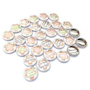 May include: A collection of 20 round, white button badges with green and orange floral designs. Each badge features a different inspirational message, such as "You are special", "You are loved", and "You are awesome".