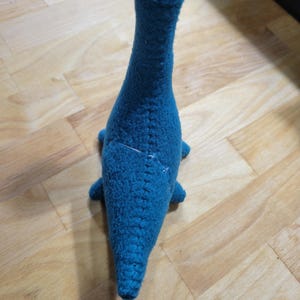 May include: A teal blue, handmade, stuffed dinosaur toy. The dinosaur has a long neck, small arms, and a tail. The toy is made of a textured fabric and is sitting on a wooden surface.