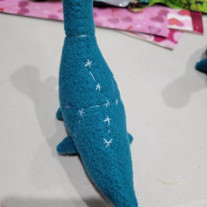 May include: A teal plush toy shaped like a sea creature, possibly a plesiosaur. The toy has white embroidered star patterns on its body. The plush has small fins and a long neck. The toy is made of a soft, textured material.