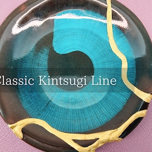 May include: A round, decorative plate with a turquoise and black design, featuring a classic Kintsugi line. The plate has a glossy finish and is accented with gold-coloured repair lines, showcasing the Japanese art of repairing broken pottery.