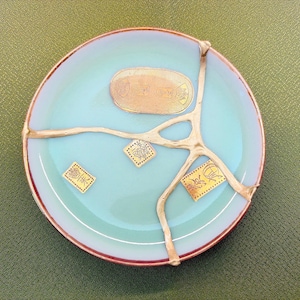 May include: A round, light blue ceramic plate with gold-colored kintsugi repair. The plate has a copper-colored rim and several gold accents. There are also gold-colored stamps with Japanese characters on the plate.