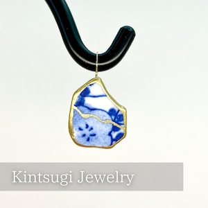May include: A Kintsugi Jewelry pendant featuring a fragment of blue and white porcelain with gold-colored accents. The porcelain has a floral pattern. The pendant is suspended from a black hook. The text "Kintsugi Jewelry" is below the pendant.