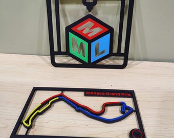 May include: Two black framed wall art pieces. The top piece features a 3D printer design with a cube displaying the letters M, L, and M in red, green, and blue. The bottom piece is a track layout with the text "Monaco Grand Prix".