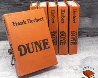 Może przedstawiać: Kolekcja pomarańczowych książek Franka Herberta, w tym "Dune", "God Emperor of Dune", "Heretics of Dune" i "Chapterhouse: Dune". Tytuły i nazwisko autora są wydrukowane na czarno. Książki są prezentowane na szarej powierzchni.