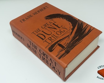 May include: A hardcover book titled "The Great Dune Trilogy" by Frank Herbert. The book has a brown cover with black lettering and a graphic illustration. The book's pages are white.