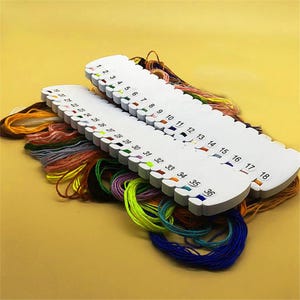Könnte beinhalten: Zwei weiße Stickgarn-Organizer aus Kunststoff mit nummerierten Schlitzen von 1 bis 36. Jeder Schlitz enthält eine andere Farbe Stickgarn, darunter Gelb-, Orange-, Rot-, Grün-, Blau-, Lila- und Brauntöne, die ordentlich in Schlaufen liegen.