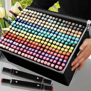 May include: A black case packed with a vibrant array of art markers, arranged in a grid pattern. The markers display a full spectrum of colours. Two black markers with coloured tips are in the foreground. The case is held by a person.