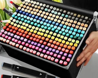 May include: A black carrying case filled with a rainbow assortment of art markers. The markers are arranged in a grid, showcasing a wide spectrum of colors. Two black markers with colored tips are in the foreground. The case is held by a person.