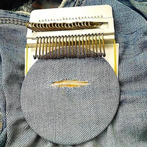 May include: A denim repair kit with a circular patch and a white plastic frame holding metal hooks. The patch is placed over a hole in a pair of blue jeans, ready for repair. The kit is designed for mending clothing.