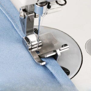 Pode incluir: Close-up do calcador e da agulha de uma máquina de costura em ação, costurando tecido azul claro. Os componentes metálicos da máquina estão em foco, destacando a precisão da costura. O tecido está sendo guiado pela máquina.