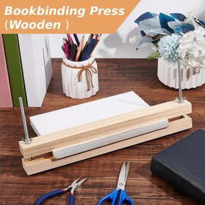 May include: A light-colored wooden bookbinding press featuring two vertical metal threaded rods with adjustable wing nuts. The press holds a stack of white paper sheets between two rectangular wooden boards, designed for securing book pages.