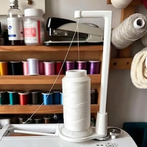 Op de afbeelding: Een witte plastic garenstandaard met een verticale arm en een horizontale bovengeleider, die een grote klos wit naaigaren vasthoudt. De draad loopt van de klos omhoog door de geleider, ontworpen voor gebruik met een naaimachine.