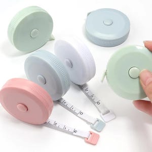 Op de afbeelding: Een verzameling van vijf ronde, intrekbare meetlinten in pasteltinten van roze, lichtblauw, wit en lichtgroen. Elke plastic behuizing is voorzien van een centrale drukknop om het witte lint met zwarte markeringen in te trekken.