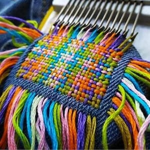 Könnte beinhalten: Nahaufnahme eines farbenfrohen Webprojekts in Arbeit. Das gewebte Quadrat zeigt eine lebendige Mischung aus orangefarbenen, grünen, blauen und violetten Fäden. Das Projekt befindet sich auf einer Jeansstoffbasis, mit farbigen Fransen, die sich von den Rändern erstrecken.