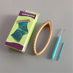 Puede incluir: Un juego de telar de punto con una caja, un telar ovalado, un ganchillo y una aguja lanera. La caja presenta una imagen de un calcetín tejido sobre un fondo verde con lunares blancos. El telar ovalado es de color beige y las herramientas tienen mangos azules.