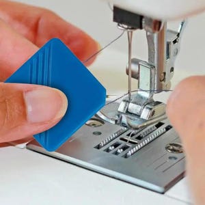 Könnte beinhalten: Ein quadratisches Nadeleinfädler-Werkzeug aus blauem Kunststoff mit drei parallelen Rillen auf der Oberfläche, positioniert neben dem Metall-Nähfuß und der Nadel einer weißen Nähmaschine mit einem dünnen schwarzen Faden.