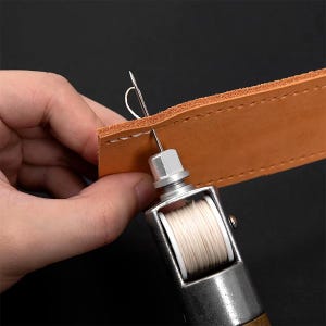 May include: A metal leather stitching awl tool with a wooden handle and a spool of white thread. The needle is shown piercing a strip of tan leather, creating a stitch with white thread. The tool features a silver metal frame and a spool.