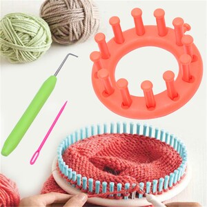 May include: A round orange knitting loom, a green handled metal hook tool, a pink plastic yarn needle, and a round white knitting loom with light blue pegs containing a partially knitted red fabric piece, alongside two balls of yarn.