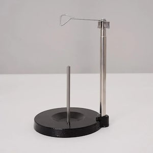 Op de afbeelding: Een metalen garenstandaard met een zware, ronde zwarte voet en een centrale verticale pin. Een hoge, gepolijste zilverkleurige stang komt vanaf de zijkant omhoog, bekroond met een horizontale draadarm voor het geleiden van het garen.