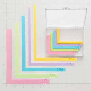 Pode incluir: Um conjunto de réguas de quilting em forma de L nas cores pastel rosa, verde-limão, azul-claro, lavanda e amarelo. Cada régua apresenta medidas em polegadas em relevo, incluindo 1/2, 3/8, 5/16, 1/4 e 3/16. Inclui um estojo transparente.