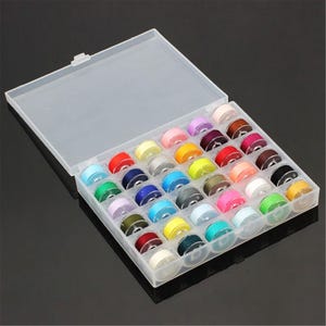 May include: A clear plastic storage box containing a selection of sewing machine bobbins in various colours. The bobbins are neatly arranged, displaying shades of red, blue, yellow, green, and purple. The box is open, allowing the contents to be seen.