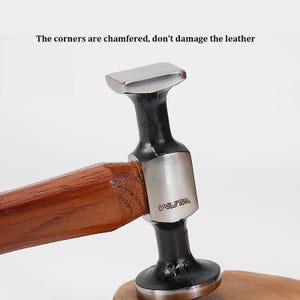 Può includere: Un martello in metallo con testa in argento lucido e manico in legno marrone scuro. La testa presenta il marchio WUTA inciso sul lato. Il testo sopra il martello recita: The corners are chamfered, don't damage the leather.