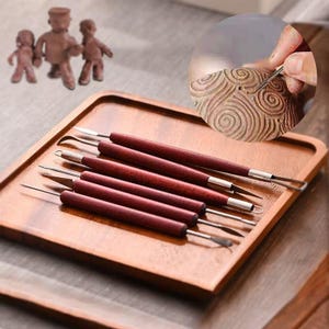 Könnte beinhalten: Ein Set aus fünf doppelseitigen Modellierwerkzeugen mit dunkel rötlich-braunen Holzgriffen und silbernen Metallspitzen. Die Werkzeuge verfügen über verschiedene Formen wie Schlaufen, Spitzen und Klingen für die Arbeit mit Ton.