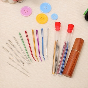 May include: A collection of sewing needles in various colors and sizes, along with several colorful buttons. The needles are arranged with some in clear tubes with red caps and others are displayed individually. A wooden needle case is also present.