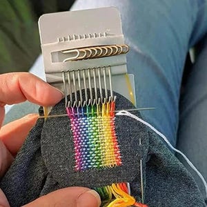 Op de afbeelding: Een kleine, grijze weefgetouw met een regenboogkleurige geweven patch in aanbouw. De weefgetouw heeft metalen tanden en een clip met goudkleurige paperclips. Een naald is in de geweven stof gestoken en kleurrijke draden zijn zichtbaar.
