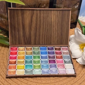 May include: A wooden watercolor paint set with a hinged lid, open to reveal an array of colorful paints. The paints are arranged in neat rows, showcasing a spectrum of hues, including reds, blues, greens, and golds. The wooden box has a natural wood grain finish.