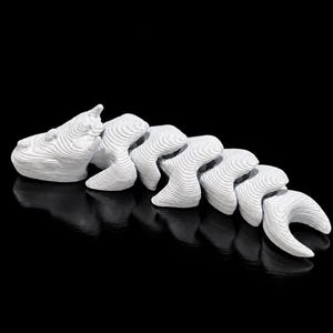 May include: A white articulated 3D printed creature figure with a segmented body and a textured, layered surface. The object features a head with two small horns and a long, flexible tail composed of several connected, curved pieces.