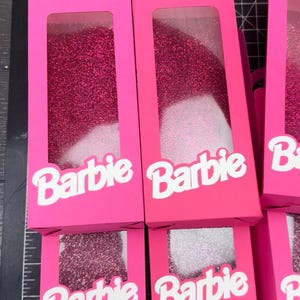 May include: Bright pink gift boxes with clear windows, filled with glitter. The boxes have a handle and the word "Barbie" in white. The boxes are arranged on a grid.