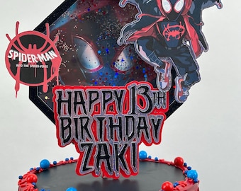 Puede incluir: Una tarta de cumpleaños decorada con el tema de Spider-Man. La tarta presenta una combinación de colores rojo y negro, con un adorno que dice "Happy 13th Birthday Zaki". La tarta está adornada con caramelos azules y rojos y una figura de Spider-Man.