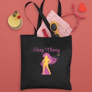 Puede incluir: Bolso tote negro con el texto "Saxy Thang" y una silueta de mujer en amarillo y rosa. La bolsa contiene gafas de sol, un labial rojo y una camisa doblada. Una bebida con una rodaja de piña y un corazón rojo también están en la imagen.