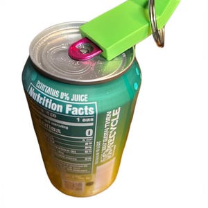 Pode incluir: Uma lata verde com um abridor de latas verde preso. O abridor tem um anel para um chaveiro. A lata tem um rótulo de informações nutricionais e as palavras "Contém 0% de suco".