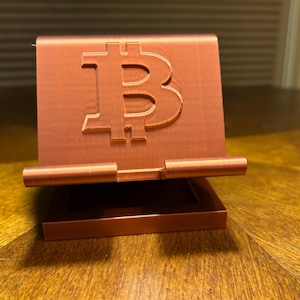 Puede incluir: Un soporte para teléfono de color cobre con un símbolo de Bitcoin. El soporte está hecho de un material sólido y tiene una base rectangular y una parte trasera con el logotipo de Bitcoin. El soporte está diseñado para sostener un teléfono.