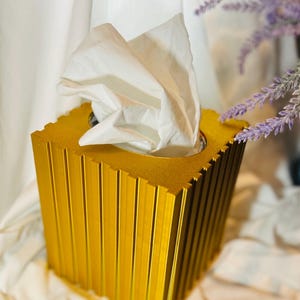 Può includere: Un porta fazzoletti dorato con un design geometrico. I fazzoletti bianchi escono dall'apertura superiore. La scatola ha linee verticali e una forma quadrata. Lo sfondo include tessuto bianco e fiori viola.