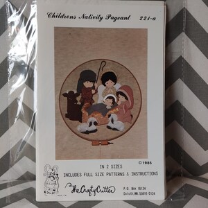 May include: A craft pattern packet titled Childrens Nativity Pageant 221-a, featuring an illustration of a nativity scene inside a circular embroidery hoop. The package includes full size patterns and instructions for two sizes, dated 1985.