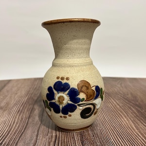 May include: A beige ceramic vase with a flared top and a round body. The vase is decorated with a blue floral design, brown leaves, and green accents. The vase is approximately 6 inches tall.